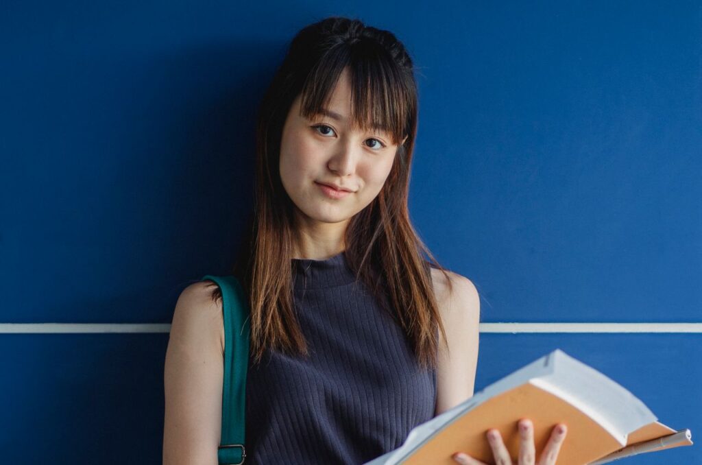 Young smiling ethnic woman with tilted head and workbooks looking at camera near tiled wall