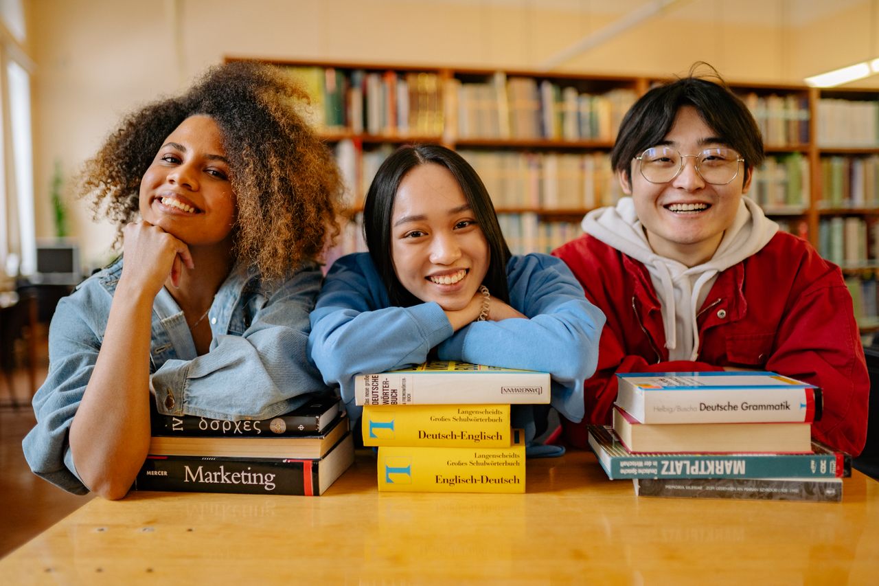 Three happy students in a library with language and marketing books.
